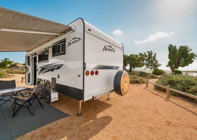 Rear view of caravan with awning extended over ground sheet and camping chairs, campsite bordered by wooden fence with trees and coastline visible in background.