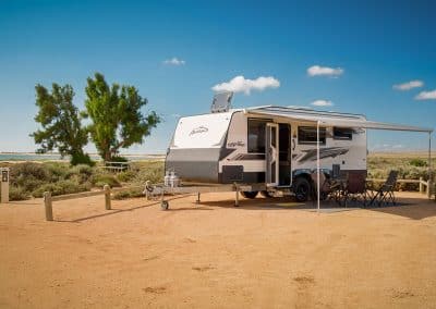 Caravan during daytime with awning extended and camping chairs and table set up underneath. Ocean and trees visible in background, sandy campsite with wooden fence.