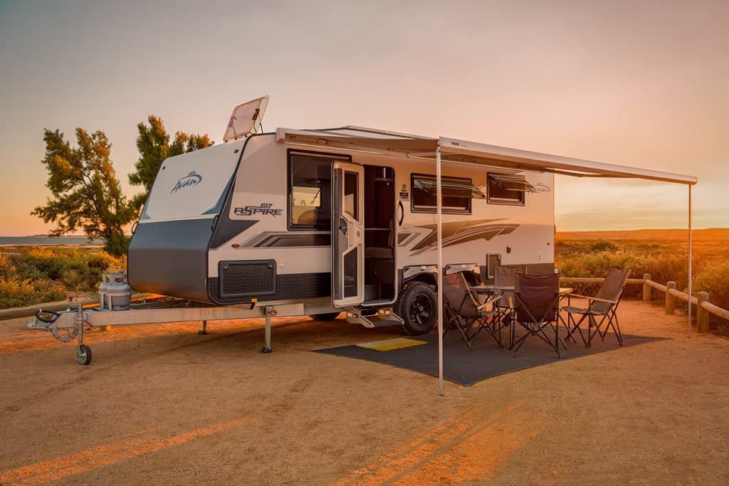 Caravan parked at sunrise campsite with awning extended, gas bottles mounted at front. Camping table and four chairs set up on ground sheet beside caravan. Sandy campsite with coastal scrubland, ocean visible in background under orange sky.