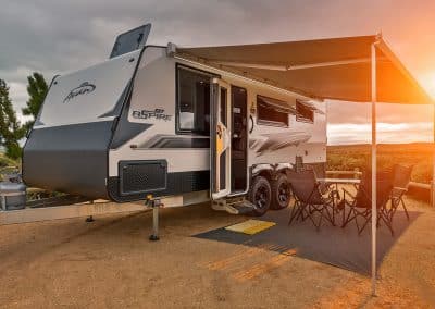 Close-up exterior view of caravan with door open, awning extended over camping table and chairs on ground sheet, gas bottles at front, sunrise glow over distant hills.