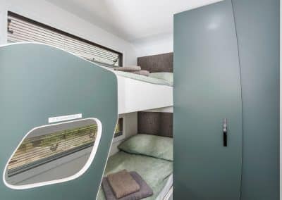 Close-up of caravan bunk beds with sage green bedding and folded grey towels, tall storage cupboard to the right, and window with flyscreen blind visible on left.