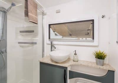 Caravan en-suite with glass-walled shower, white vessel sink on stone-look benchtop, soap dispenser and small potted plant. Large window with block-out blind above sink, grey towel hanging by shower.