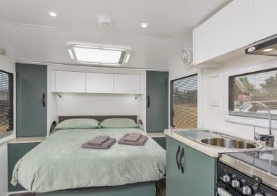 Front-facing view of caravan bed with sage green bedding and folded grey towels. Green wardrobe cabinets either side of bed. Kitchen counter, sink and cooktop visible on the right. Windows with blinds on both sides.