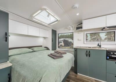 Caravan bedroom with sage green bedding on island double bed, grey towels folded on top. White overhead cabinetry, skylight above bed, window views of trees and parked vehicle. Kitchen with sink and cooktop visible in background.