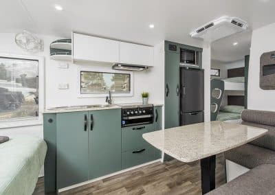 Caravan kitchen with sage green cabinetry, stone-look benchtop, gas cooktop and black fridge/freezer. Dining table with grey bench seating in foreground. Bunk beds with striped bedding visible behind partition. Timber-look flooring.