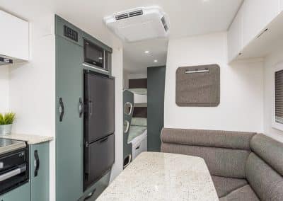 Caravan interior showing grey L-shaped dining bench with stone-look table, sage green pull-out pantry cabinet, black fridge/freezer and microwave. Bunk beds visible through partition at rear, air conditioning unit mounted on ceiling.
