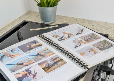 Open spiral-bound instruction folder on a caravan countertop, showing step-by-step photo guides for awning setup with labelled hands demonstrating each step. Potted plant in background.
